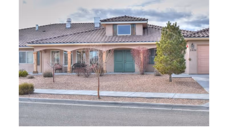 Front exterior of a single-story stucco home with a tiled roof, double green front doors, gravel landscaping, and a small tree.
