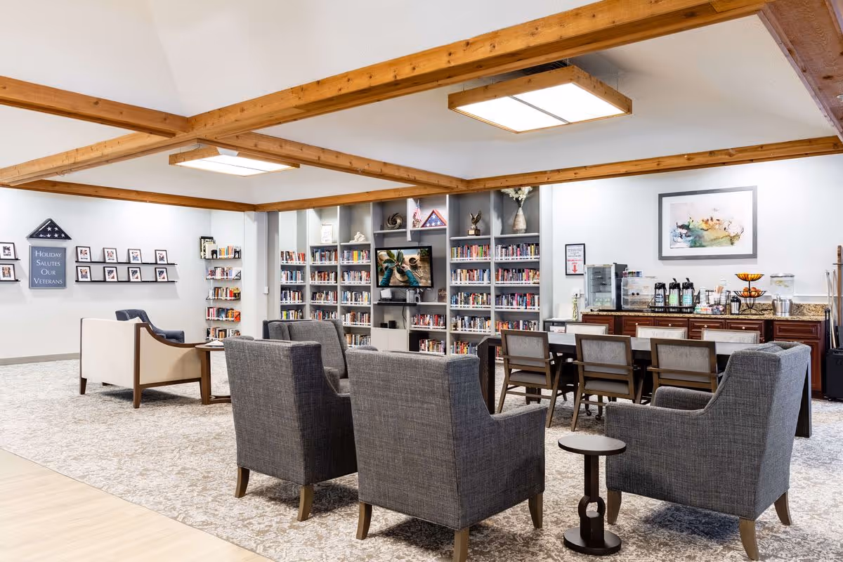 A spacious and well-lit common area with wooden ceiling beams, featuring a seating arrangement of gray armchairs and a beige sofa around a small table. In the background, there is a large bookshelf filled with books and a TV mounted in the center. To the right, a long table with chairs is set up near a counter with coffee and beverage dispensers. The walls are decorated with framed pictures and a sign that reads 'Holiday Salutes Our Veterans.'