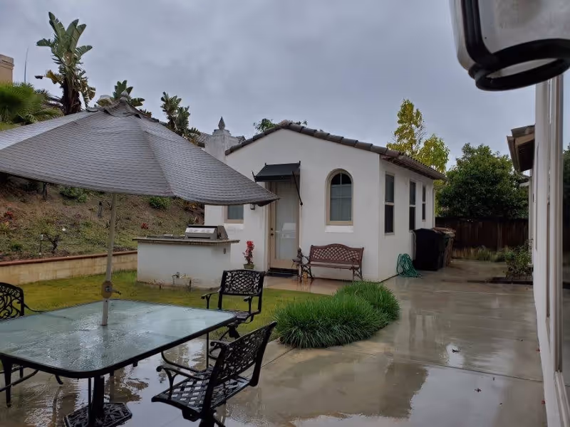 Rainy backyard patio with a glass table and umbrella, metal chairs, a small white guest house, bench, and outdoor grill.