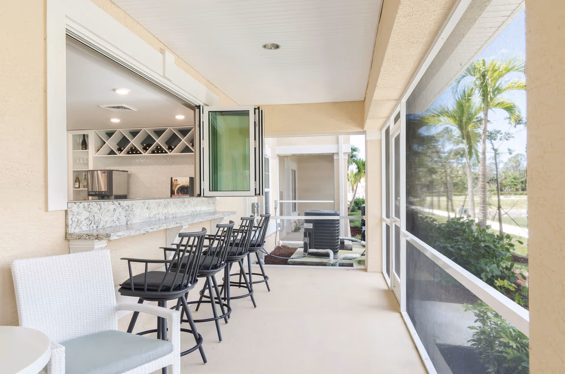 A covered outdoor patio area with a marble countertop bar and several black bar stools. There is a white chair with a cushion in the foreground and a window opening to an indoor space with wine racks and bottles. The patio is screened with a view of palm trees and greenery outside.