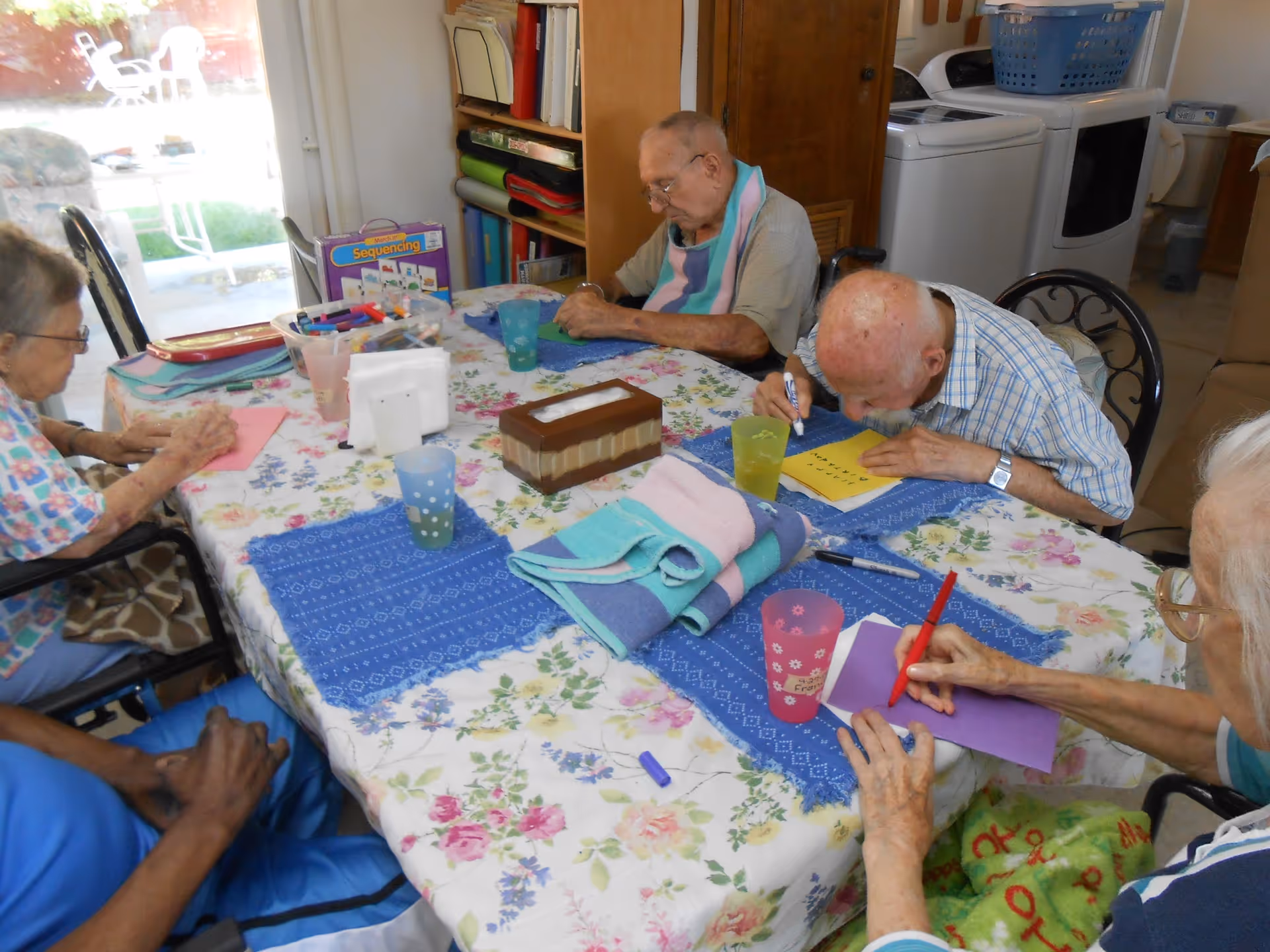 A group of elderly individuals sitting around a table engaged in a craft activity, writing or drawing on colorful paper. The table is covered with a floral tablecloth and blue placemats, with various craft supplies and cups placed on it. The room appears to be a cozy indoor setting with shelves and household items in the background.