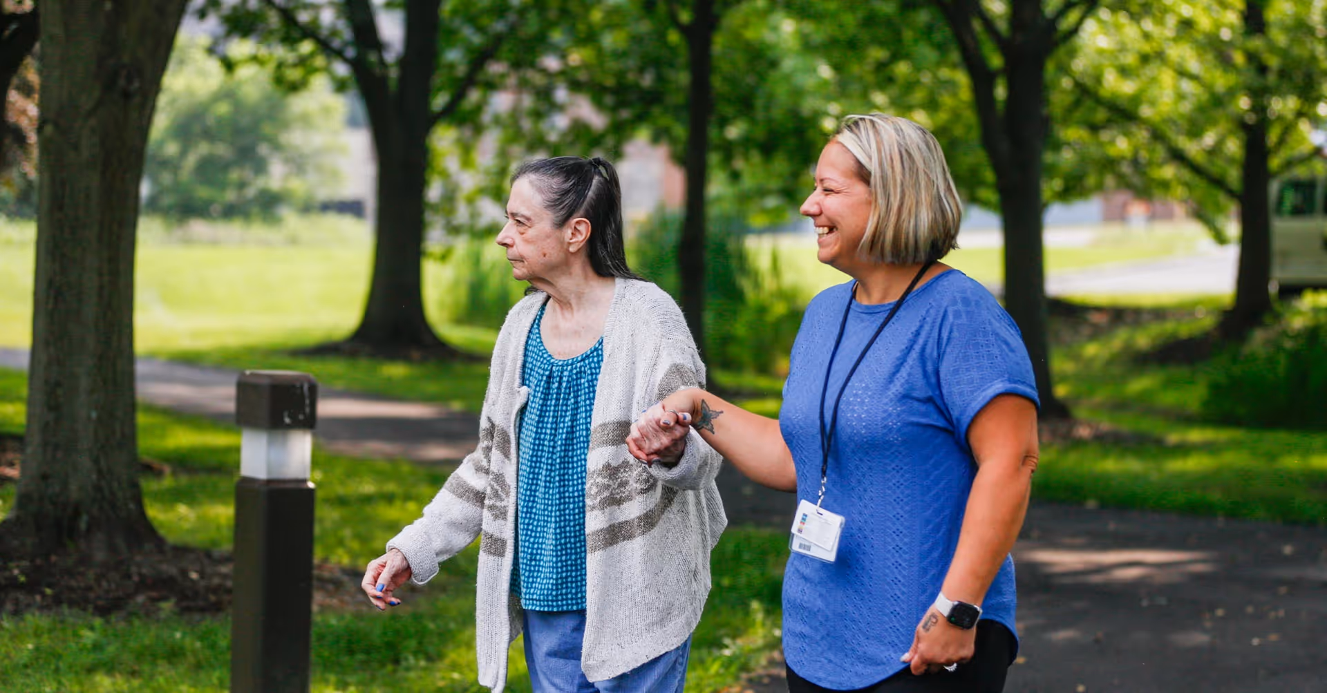 An elderly woman and a caregiver holding hands and walking outside on a paved path surrounded by green grass and trees. The caregiver is smiling and wearing a blue shirt with a name badge, while the elderly woman is wearing a blue top and a beige cardigan.