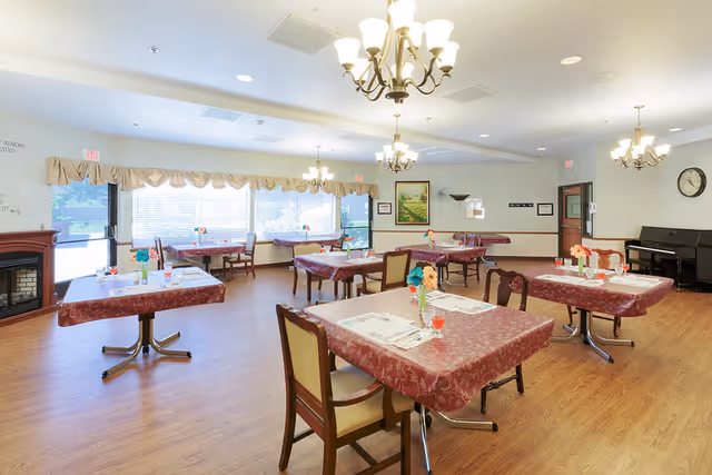 A bright dining room with several tables covered in red patterned tablecloths, each set with placemats, glasses, and small flower arrangements. The room has wooden flooring, large windows with blinds and valances, chandeliers hanging from the ceiling, a wall clock, a piano, and framed artwork on the walls.