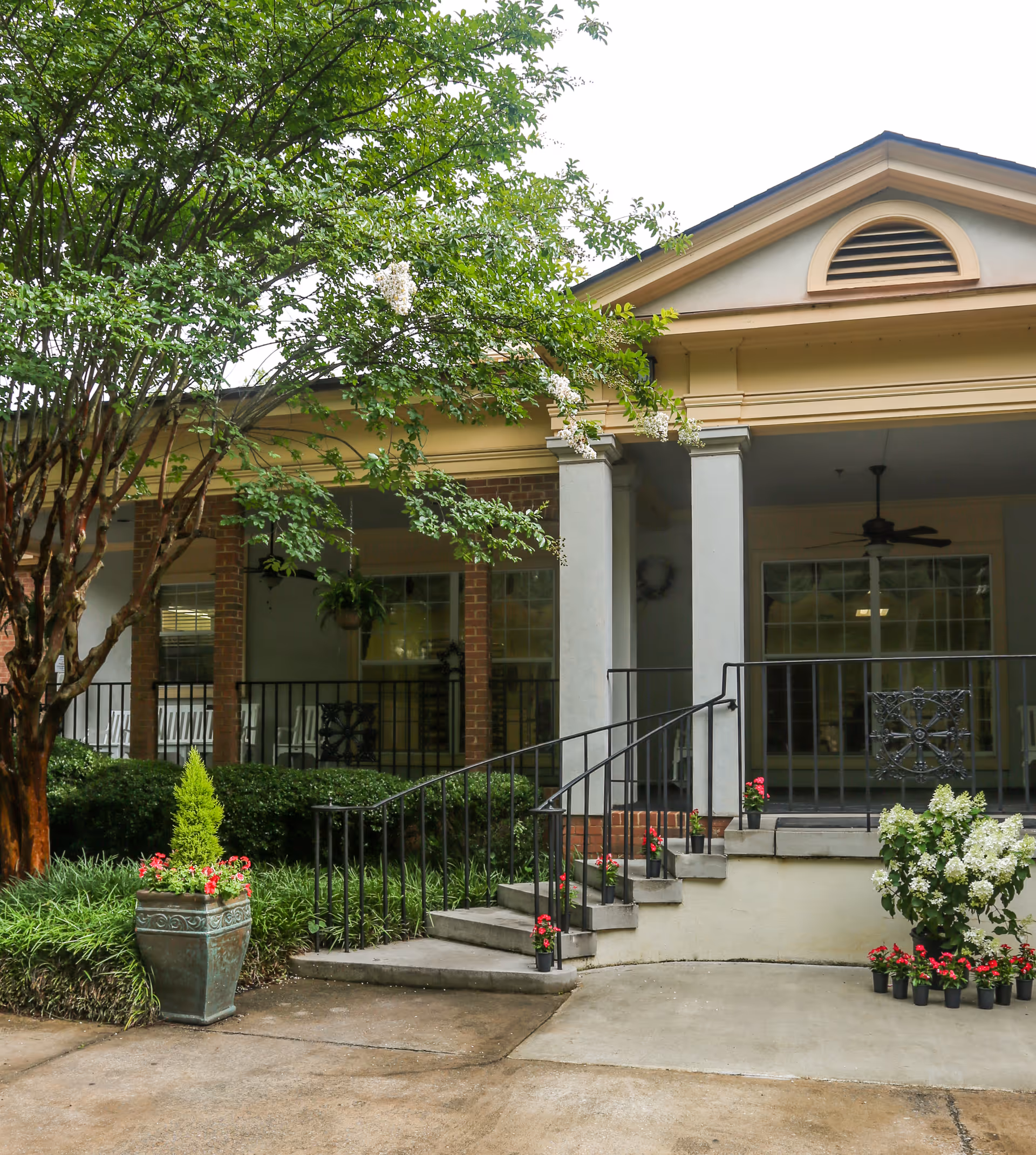 Entrance to a building with a small staircase and black metal railing, surrounded by greenery and potted flowers. The building has brick and light-colored walls with large windows and a covered porch area with ceiling fans.