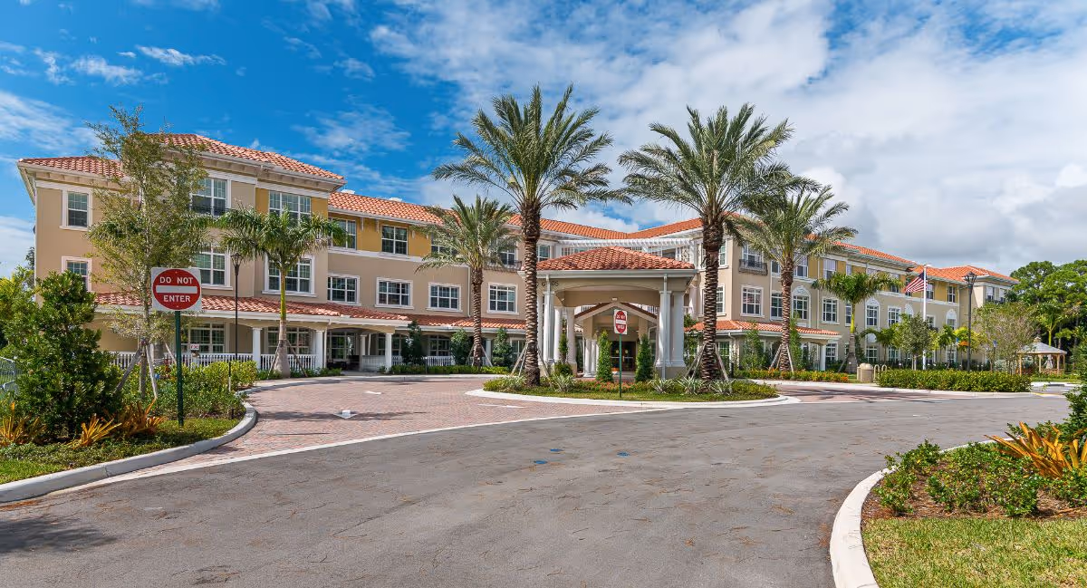 Exterior view of The Barclay at Boynton Beach, a multi-story senior living facility with a beige and yellow facade, red-tiled roof, and multiple windows. The entrance features a covered driveway with columns and is surrounded by palm trees and landscaped greenery under a partly cloudy blue sky.