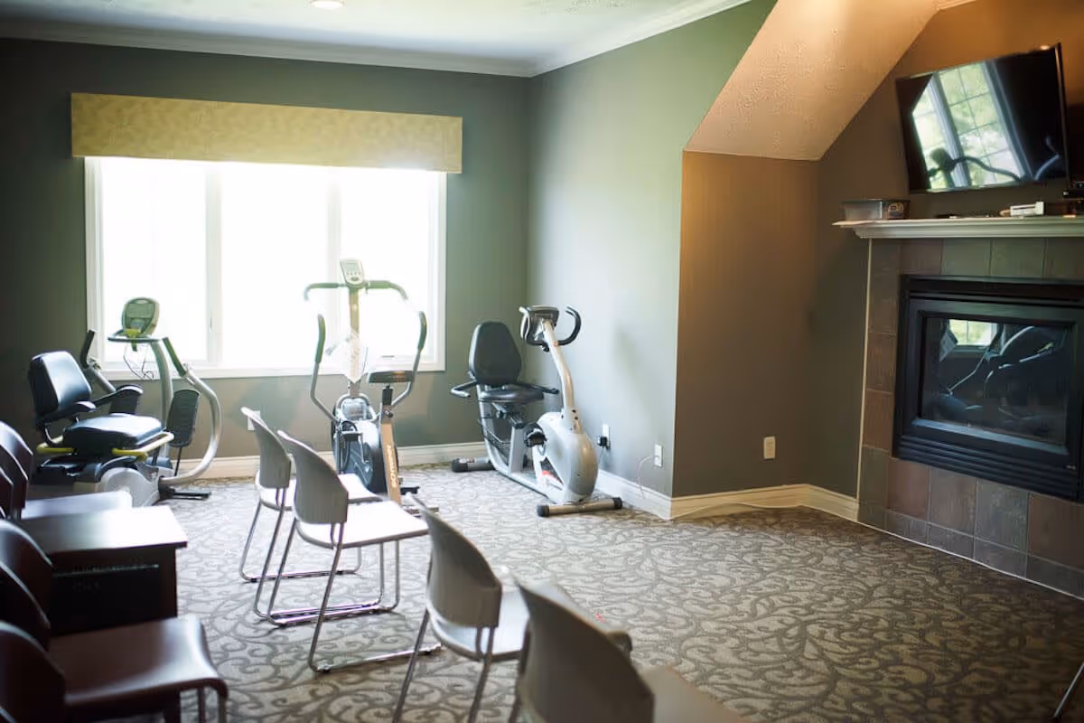 A small exercise room with three stationary exercise bikes positioned near a large window with a beige valance. The room has patterned carpet flooring, gray walls, a fireplace with a mounted flat-screen TV above it, and several chairs arranged in rows facing the exercise bikes.