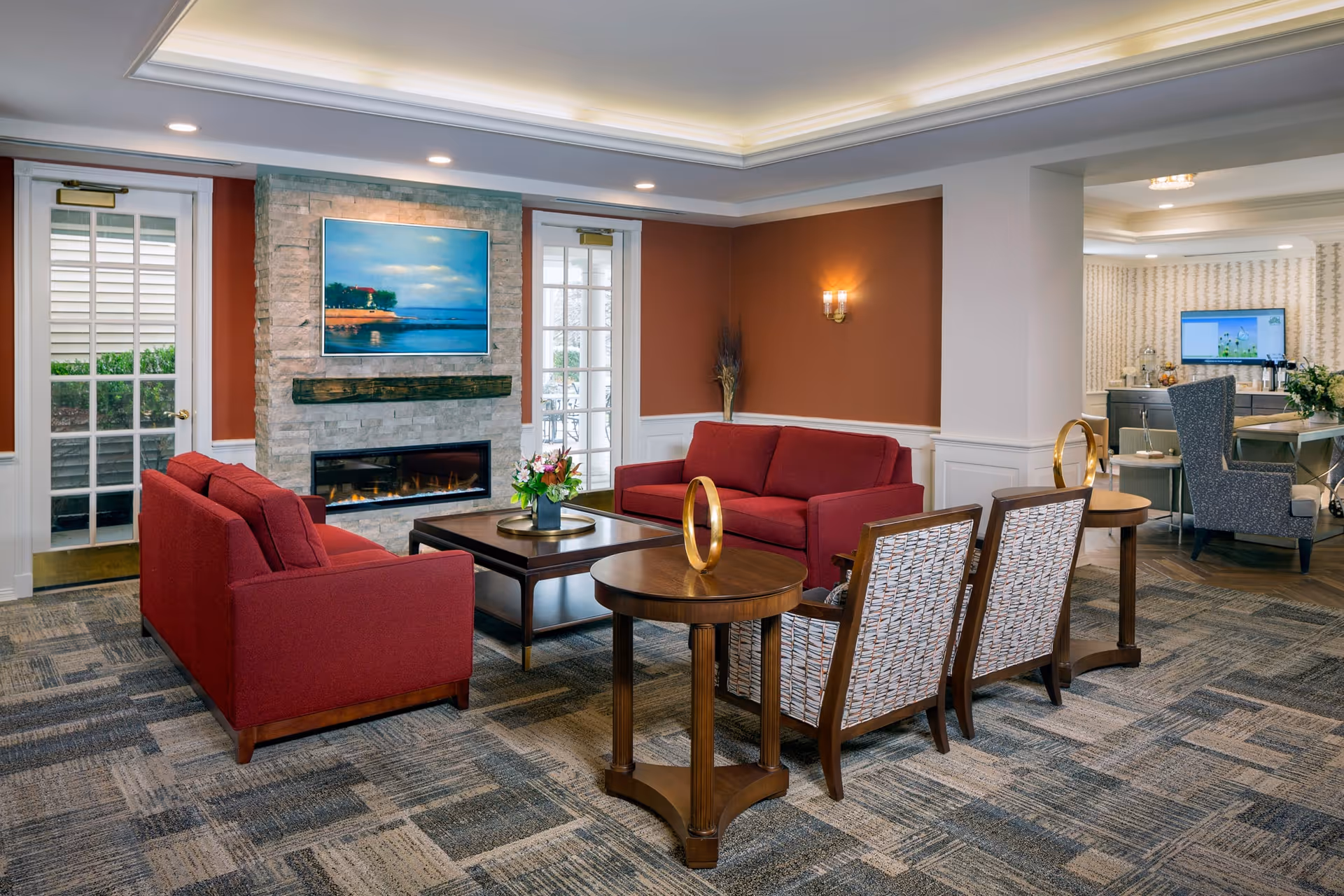 A cozy living room area with two red sofas facing a stone fireplace with a landscape painting above it. There are two patterned armchairs and wooden side tables with decorative golden rings. The room has warm lighting, carpeted floors, and a view into an adjacent dining or common area with a television and additional seating.