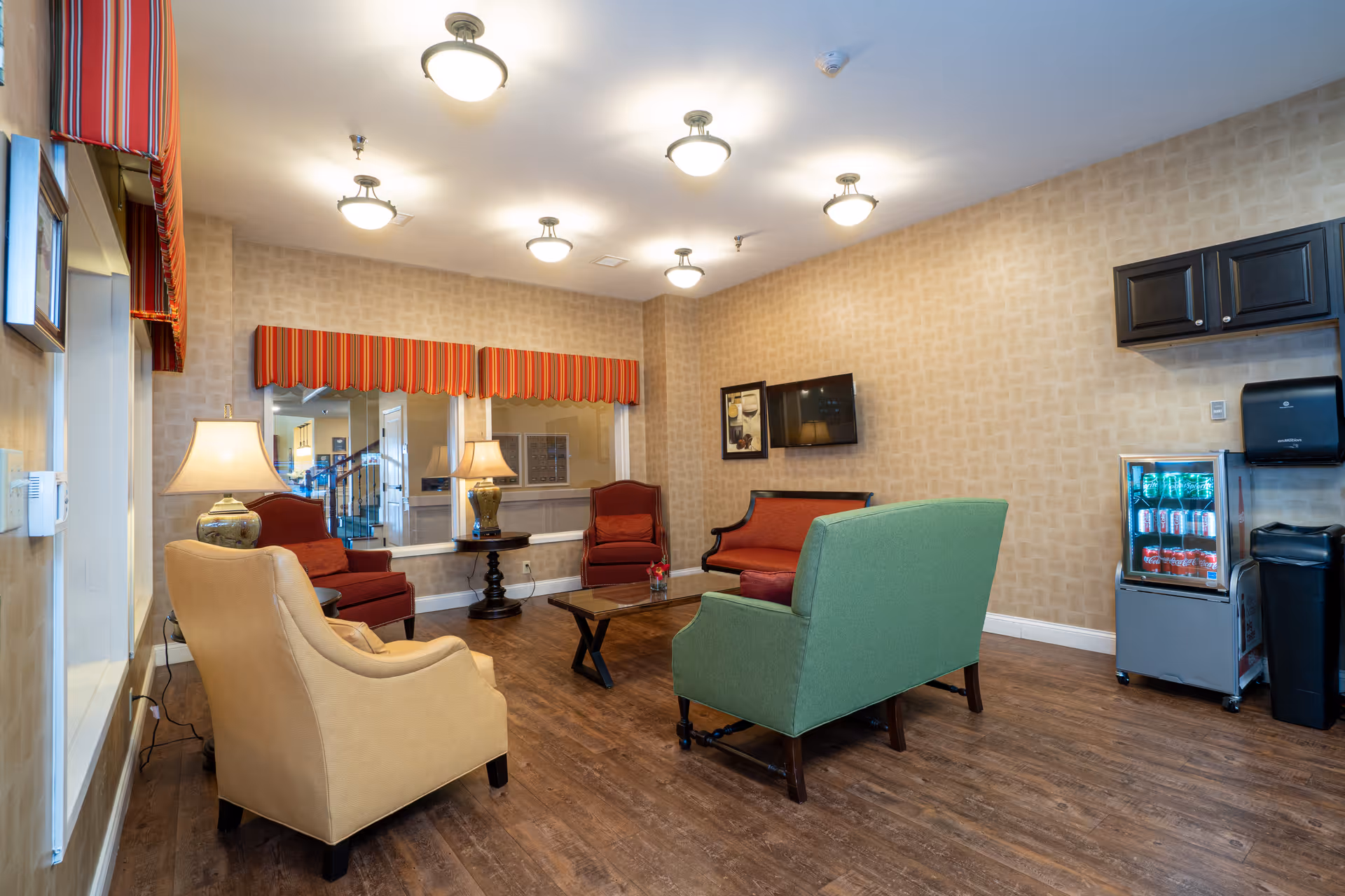 Comfortable common lounge with upholstered chairs and sofas, a coffee table, lamps, a wall-mounted TV, and a small refrigerated drinks case.