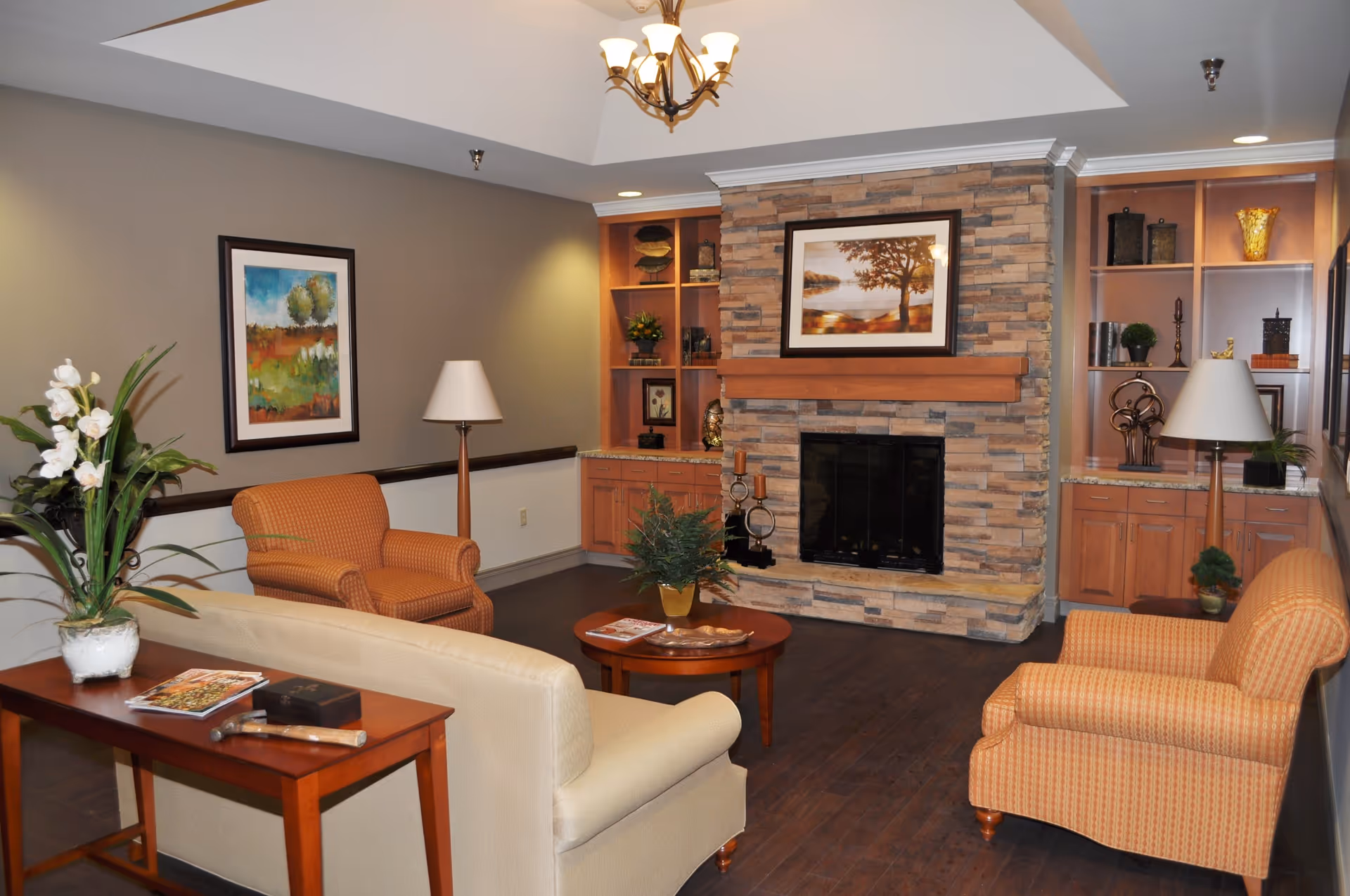 A cozy living room with a stone fireplace at the center, flanked by built-in wooden shelves and cabinets. The room features two orange patterned armchairs, a beige sofa, a wooden coffee table with a plant, and a side table with magazines and a decorative item. The walls are painted in neutral tones and decorated with framed artwork. A ceiling light fixture hangs above.