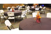 Event/dining room with round tables draped in dark tablecloths, white folding chairs, and floral centerpieces.