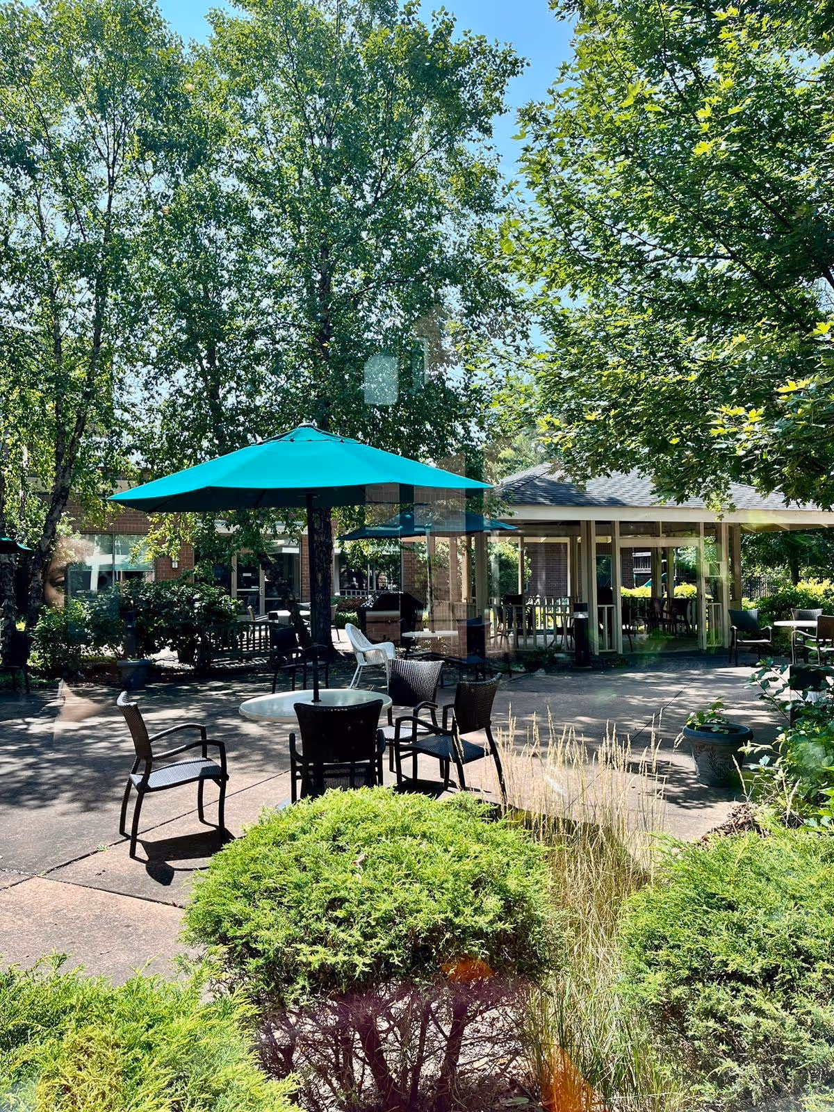 Outdoor patio area with a round table and several chairs under a large teal umbrella surrounded by green trees and bushes. In the background, there is a covered seating area with more chairs and tables.