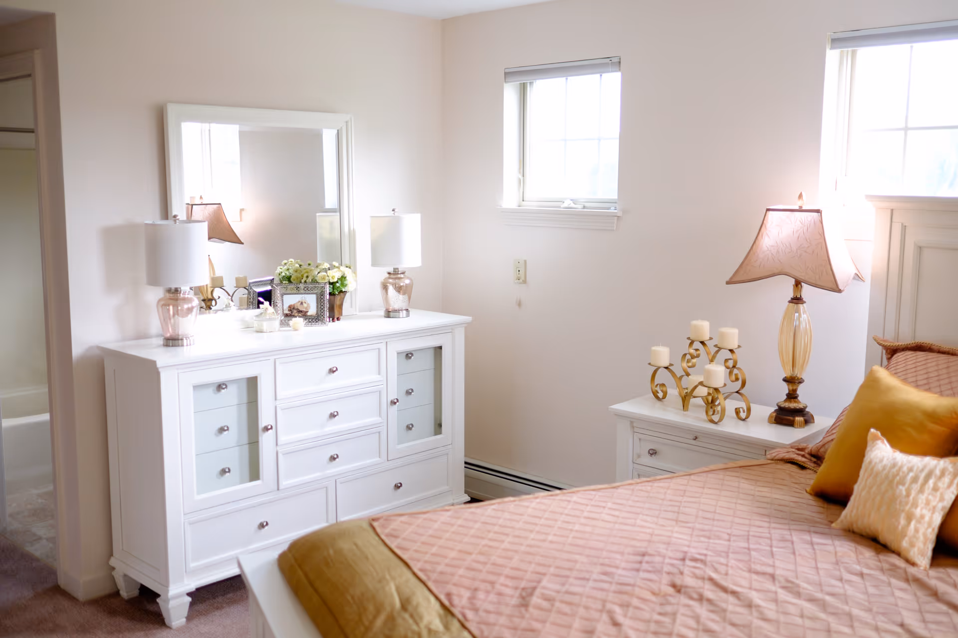 Bright bedroom with a white dresser and mirror, bedside tables and lamps, and a bed with a pink quilt and accent pillows.