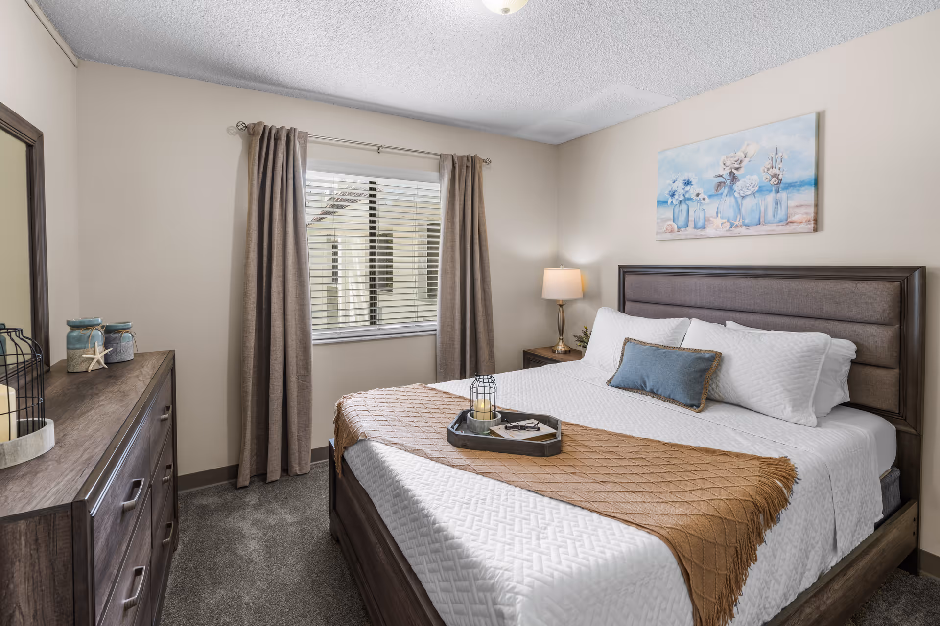 A neatly arranged bedroom with a large bed featuring white bedding, a brown throw blanket, and decorative pillows. There is a wooden dresser with decorative items on the left side, a window with beige curtains in the center, and a nightstand with a lamp on the right. Above the bed hangs a painting of blue vases with flowers.