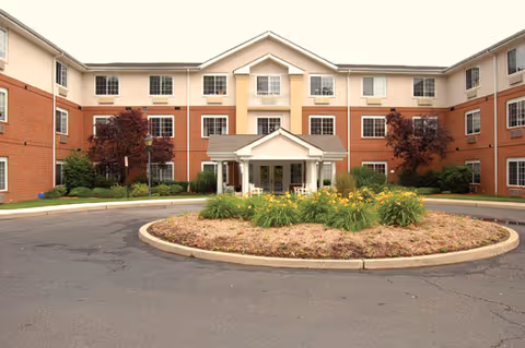 Front exterior view of a three-story senior living facility building with a circular driveway and landscaped roundabout featuring plants and flowers. The building has a brick lower facade and light-colored upper walls with multiple windows and a covered entrance.