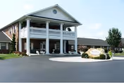 Front exterior view of a two-story brick building with white columns and a balcony, surrounded by a paved driveway and landscaped bushes. A sign near the entrance reads 'Heritage Corner Health Care'.
