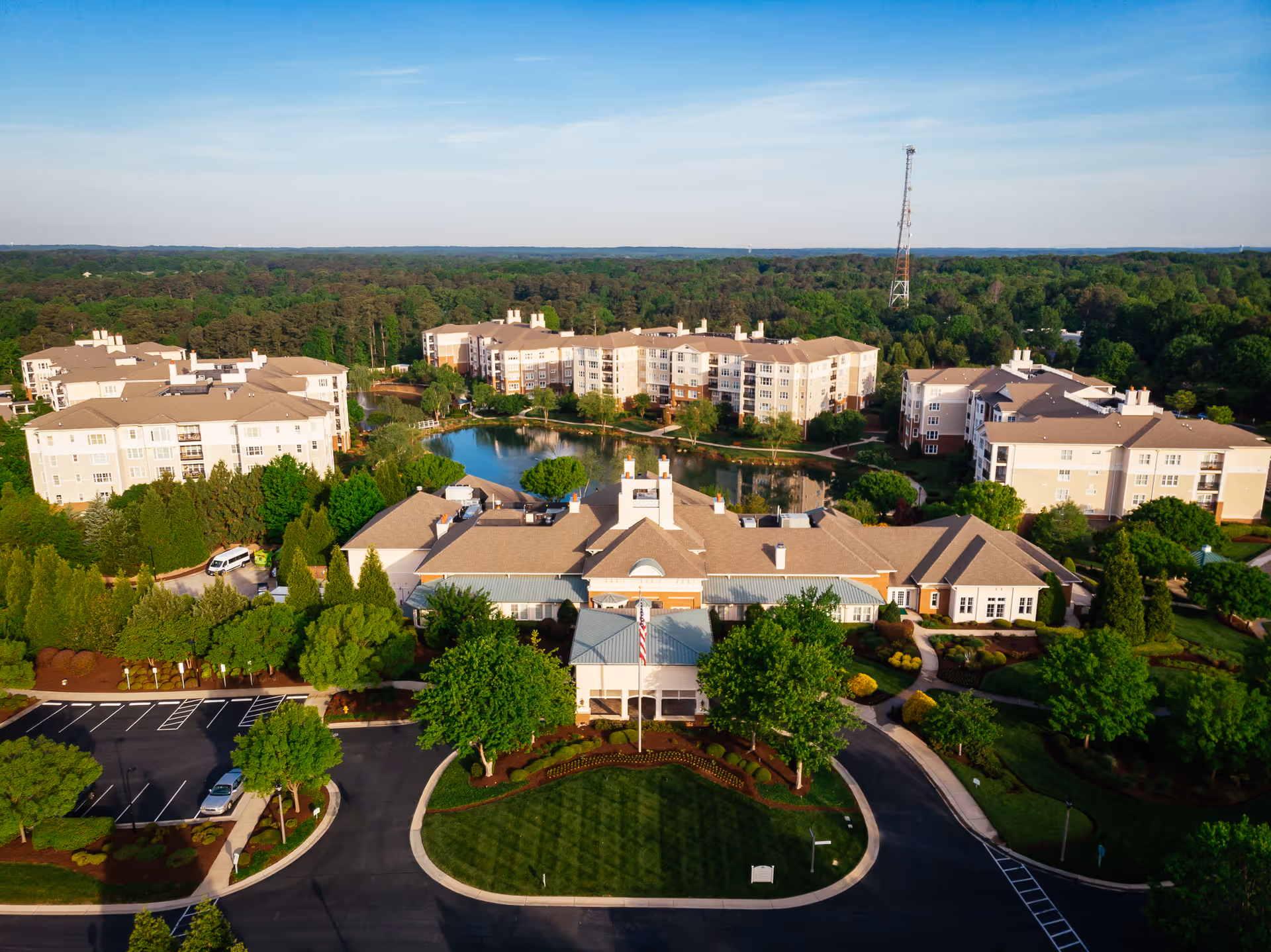 Aerial view of a landscaped senior living campus with multiple beige apartment buildings around a central pond and entrance drive.