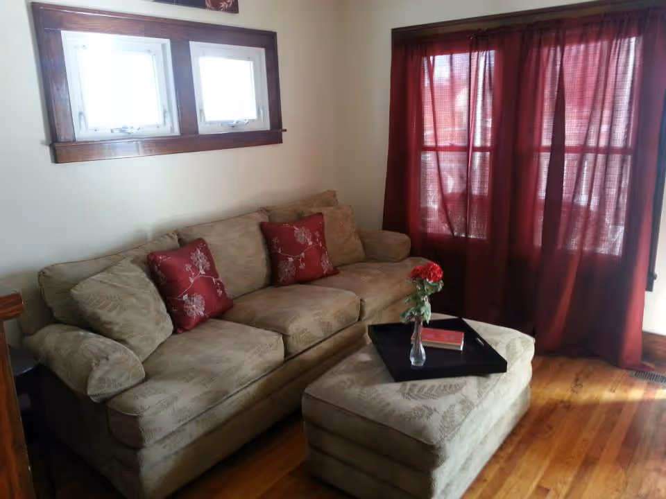 A cozy living room with a beige patterned sofa adorned with red floral cushions, a matching ottoman with a black tray holding a small vase with red flowers and a book, wooden floor, two small windows with wooden frames, and a large window covered with red curtains.