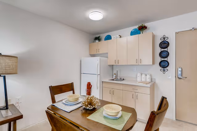 Dining area with a wooden table set for two and a compact kitchenette with refrigerator and cabinets.