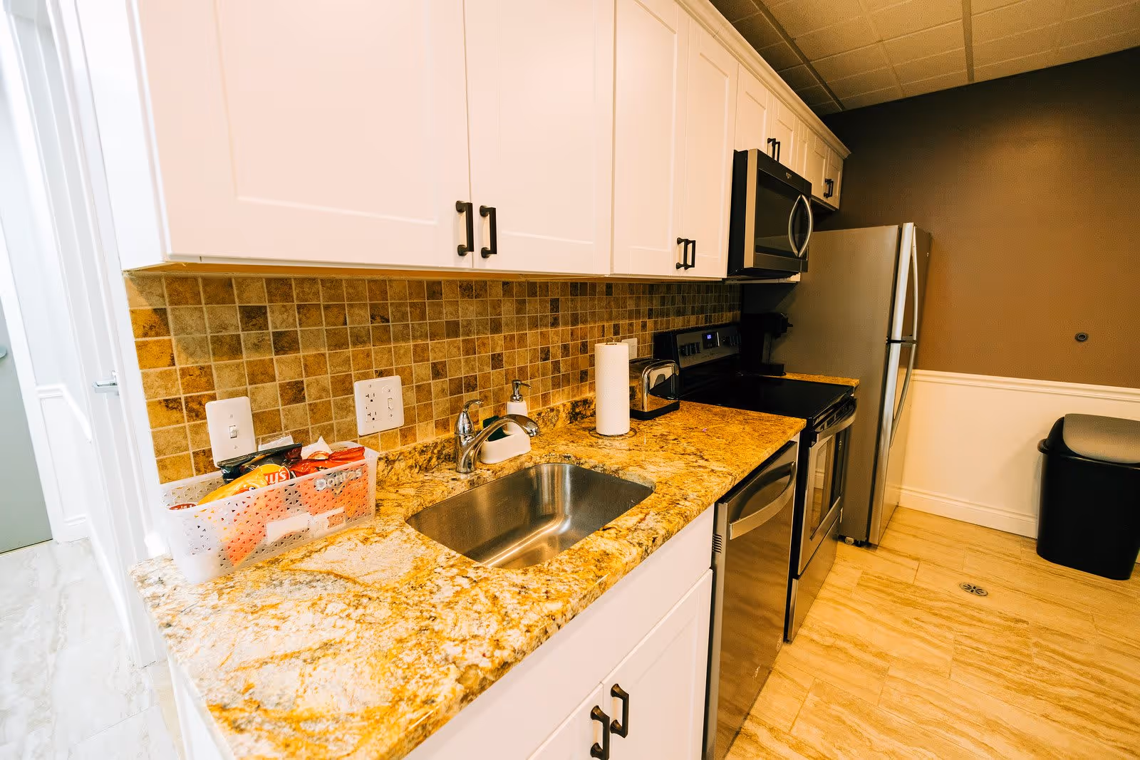 A modern kitchen with white cabinets, a granite countertop, a stainless steel sink, and stainless steel appliances including a refrigerator, stove, microwave, and dishwasher. There is a paper towel holder and a small basket with snacks on the counter. The backsplash is made of small square tiles in earth tones, and the floor is tiled in a light beige color.
