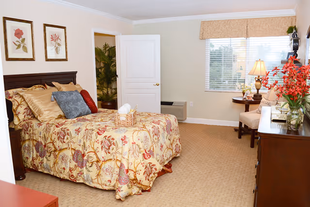 A cozy bedroom in a senior living facility featuring a bed with floral patterned bedding and multiple pillows. The room has beige carpet, light-colored walls, and two framed floral prints above the bed. There is a window with blinds and a valance, a small round table with a lamp and chair, a dresser with a vase of red flowers, and an open door revealing a plant in the adjacent room.