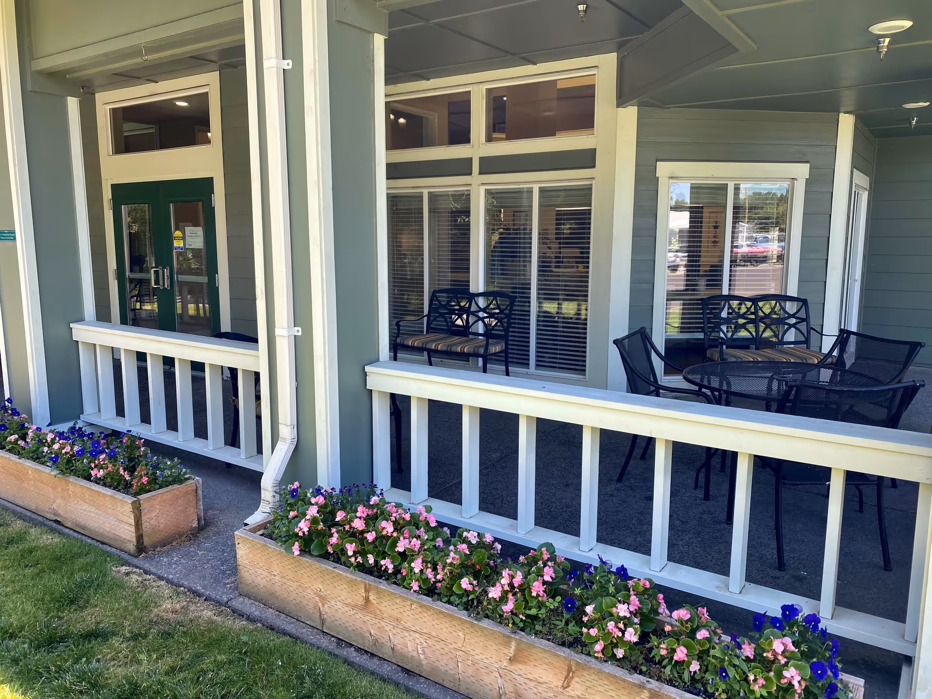 Outdoor covered patio area at Cascade Valley Assisted Living and Memory Care with a seating arrangement including a round table and chairs, two benches, and flower beds with pink and purple flowers along the edge of the patio.