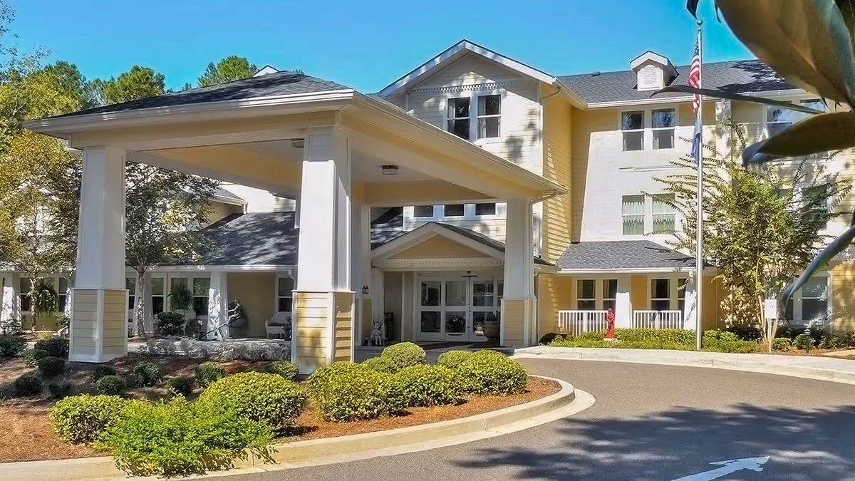 Front exterior view of a senior living facility named Ashley Park by Barclay House, featuring a covered entrance with white pillars, manicured bushes, and an American flag on a flagpole.
