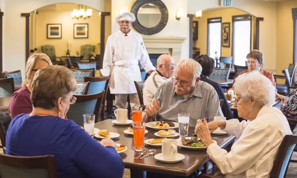 A group of elderly people sitting around a dining table enjoying a meal together in a well-lit dining room. A chef in white uniform and hat stands in the background near a fireplace and a round mirror on the wall. The room has wooden chairs and tables, with framed pictures on the walls and an exit sign visible.