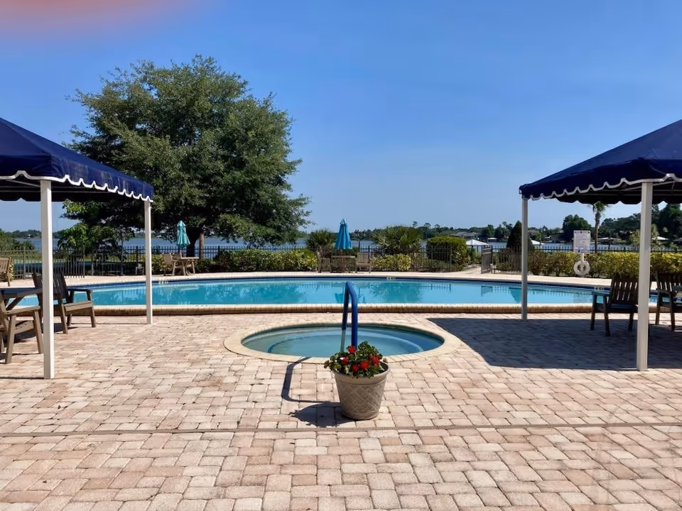 Outdoor swimming pool area with a small round hot tub in the foreground, surrounded by a paved deck. Two blue canopies provide shade over seating areas with chairs. In the background, there are trees, shrubs, and a body of water under a clear blue sky.