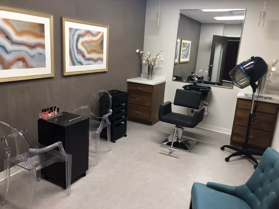 Interior salon room with a styling chair and shampoo sink, hooded hair dryer, acrylic guest chairs, a small nail station and framed artwork.