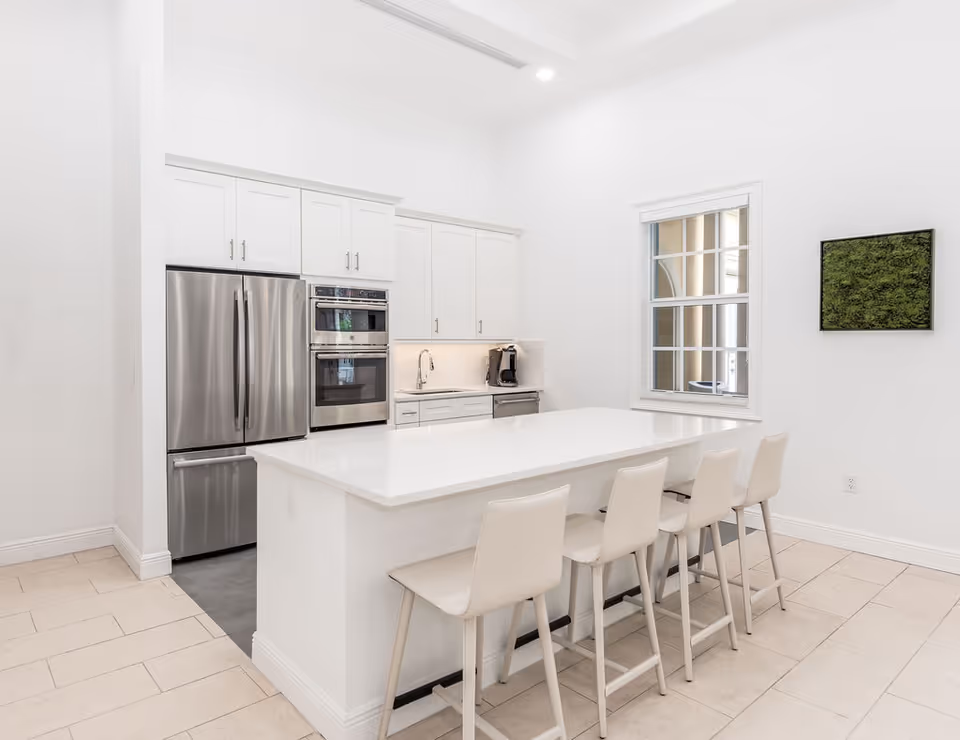 Bright contemporary white kitchen with a large island and four bar stools, stainless steel refrigerator and double oven.