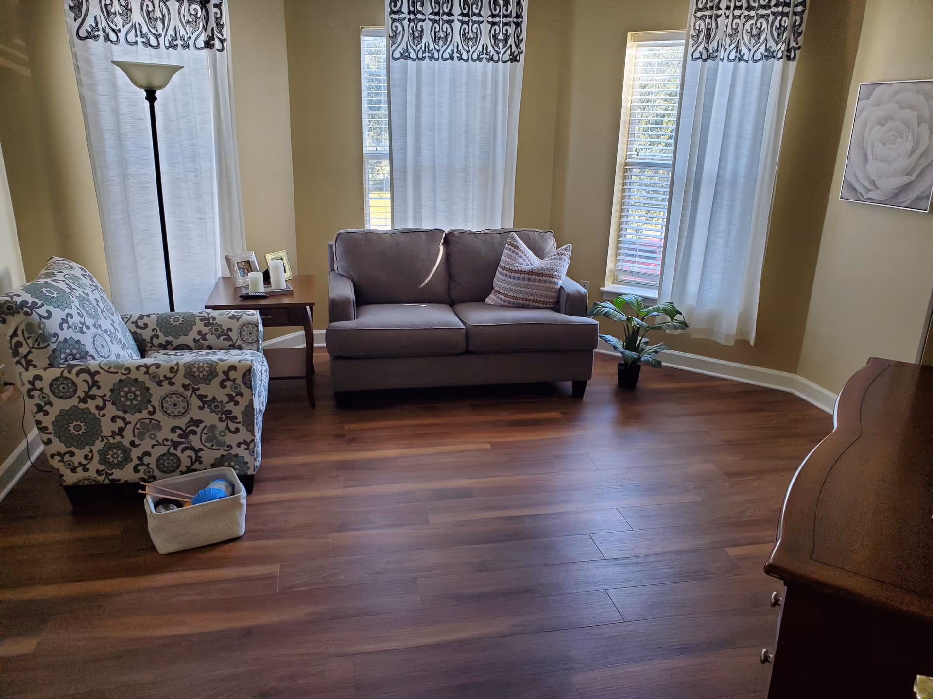 A cozy living room with wooden flooring, featuring a gray loveseat with a patterned pillow, a floral-patterned armchair, a wooden side table with picture frames and candles, a tall floor lamp, a small potted plant near the window, and three windows with white curtains that have a decorative black pattern at the top.