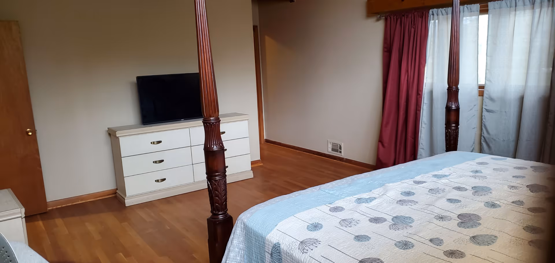 A bedroom with a wooden four-poster bed covered with a light blue and white patterned quilt. There is a white dresser with six drawers and a flat-screen TV on top against the wall. The room has wooden flooring, beige walls, a wooden door, and windows with red and white curtains.