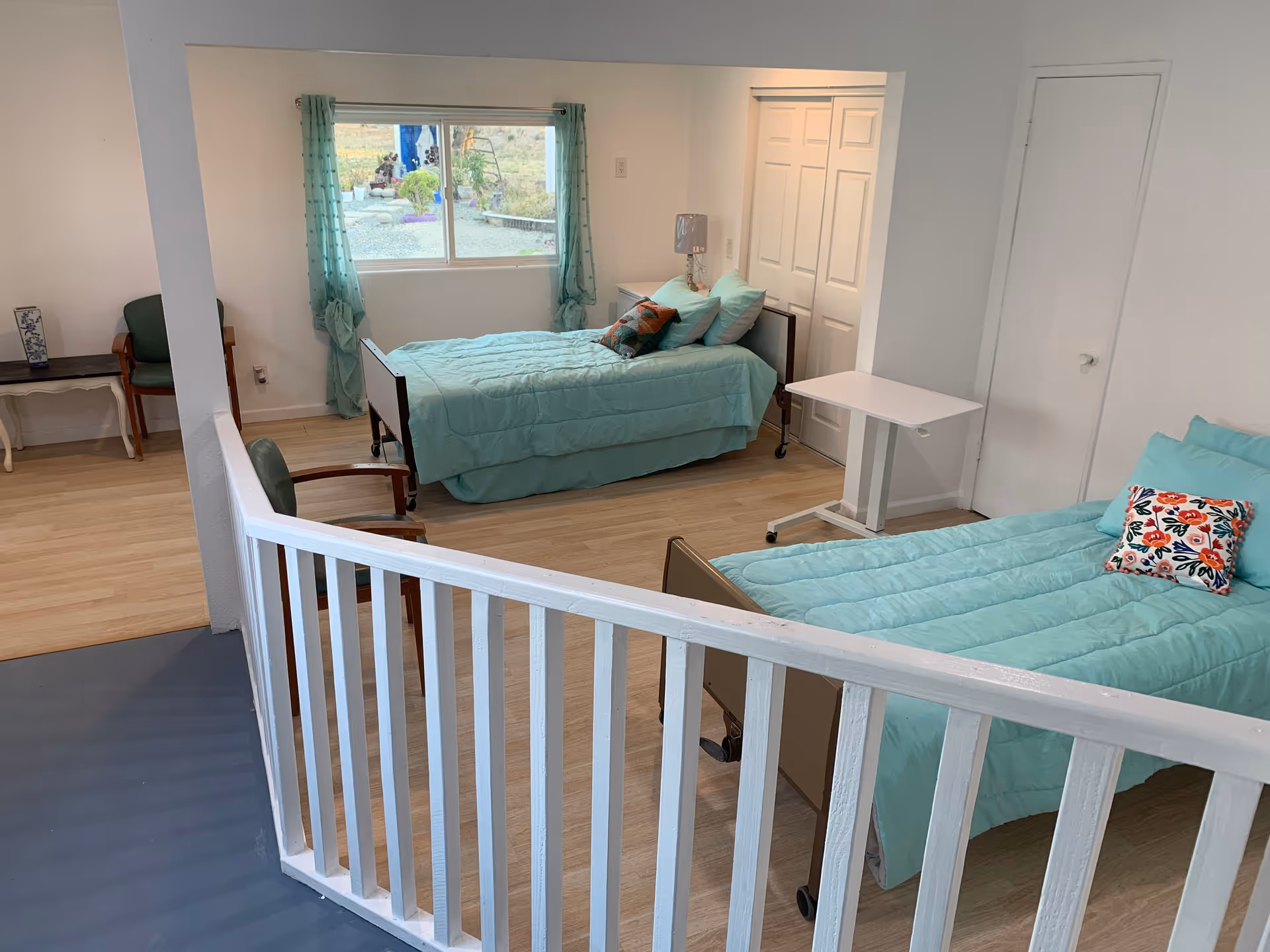 A bright room with two single beds covered in light turquoise bedding, each with decorative pillows. There is a window with sheer turquoise curtains letting in natural light. The room has light wood flooring, a white railing in the foreground, a small white adjustable table between the beds, and a couple of chairs and a small table against the wall.