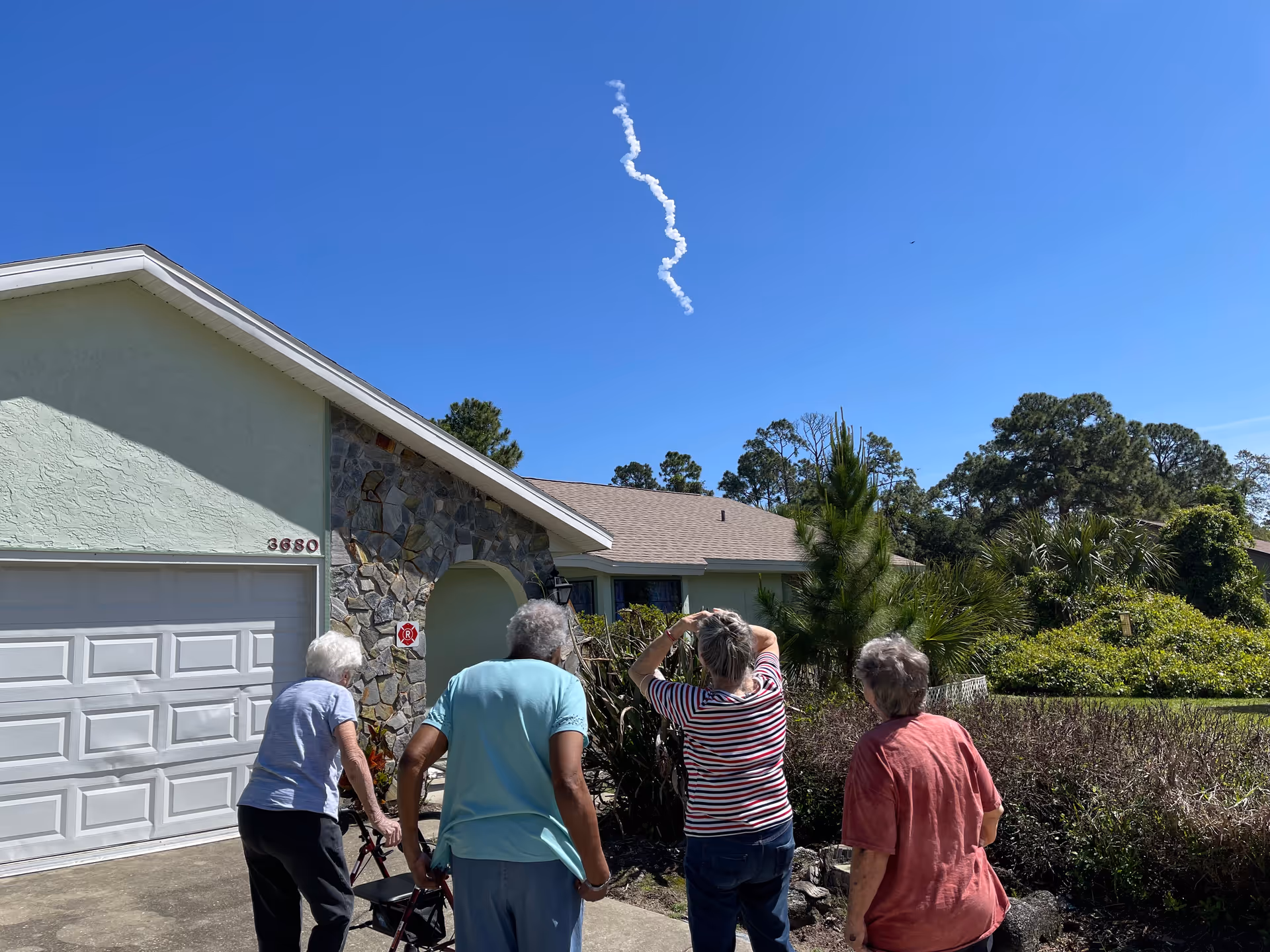 Four elderly individuals standing outside a residential home looking up at the sky where a white, curly smoke trail is visible against a clear blue sky. The house has a stone facade and a white garage door with the number 3680 on it. There are trees and bushes surrounding the house.