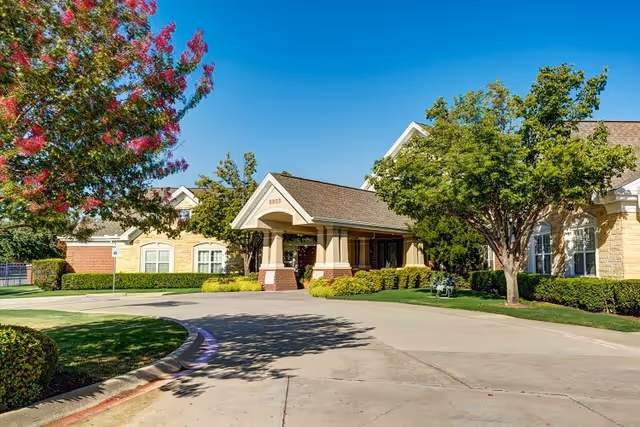 Exterior view of a senior living facility building with a covered entrance, surrounded by trees and well-maintained landscaping under a clear blue sky.
