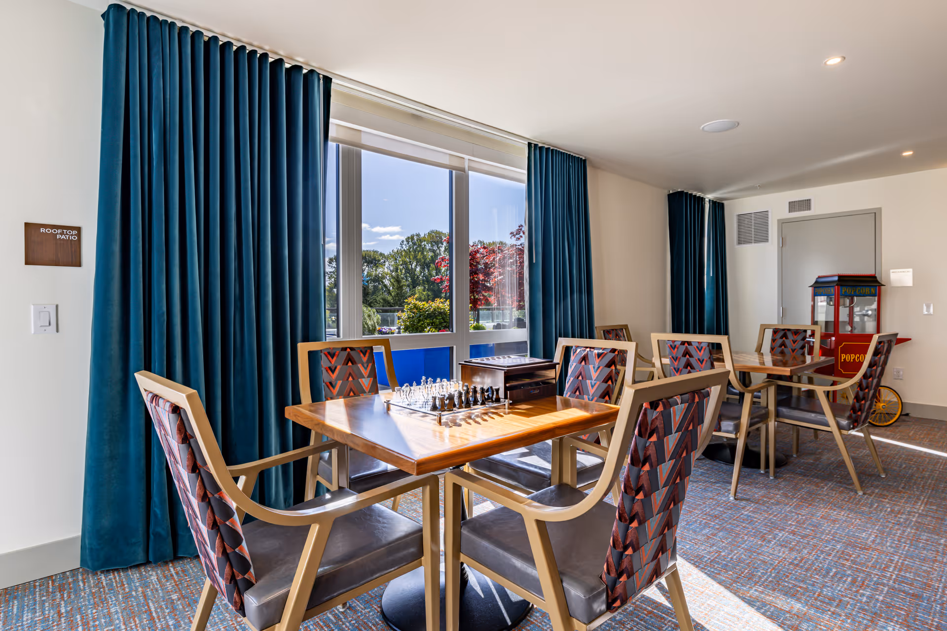 Sunlit community activity room with wooden tables and patterned chairs, a chess set on a table, and a popcorn machine in the back.