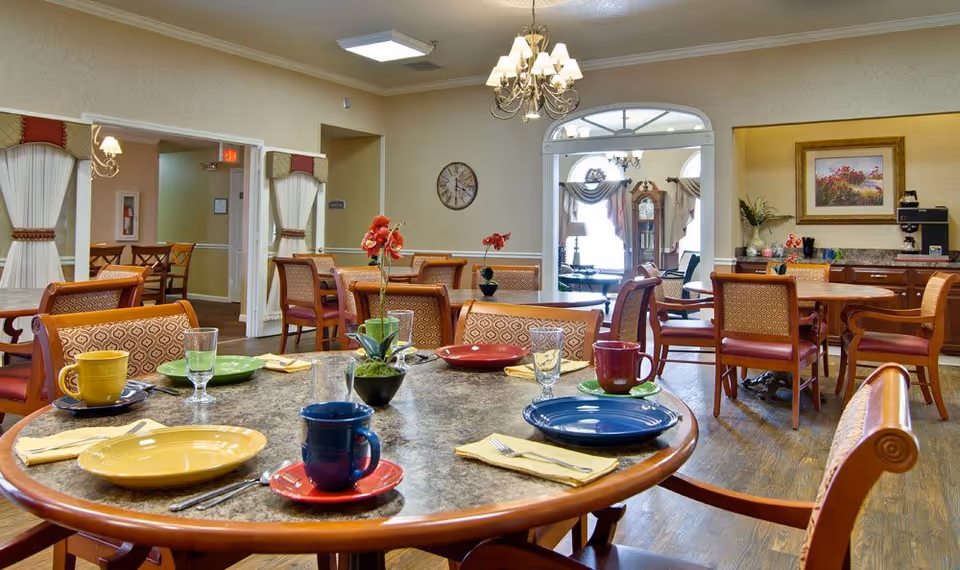 A dining room in a senior living facility with multiple round tables set with colorful plates, cups, and napkins. The room features wooden chairs with patterned upholstery, a chandelier, wall clock, and a coffee station in the background. There is an open doorway leading to another room with a grandfather clock and additional seating.