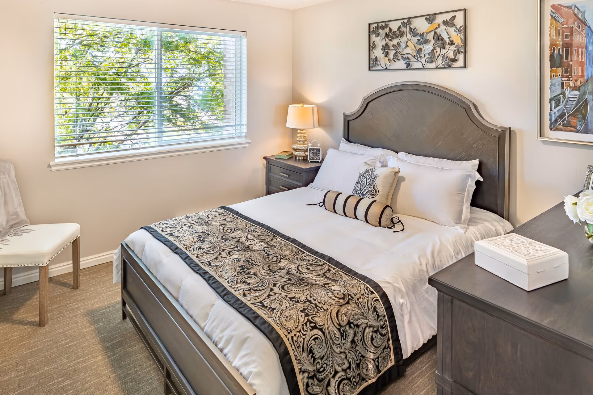 A neatly made bed with white linens and decorative pillows in a bedroom. The bed has a dark wooden headboard and a patterned throw blanket. There is a nightstand with a lamp, clock, and book next to the bed. A window with blinds lets in natural light, and there are framed artworks on the walls. A chair with a light-colored throw is placed near the window.