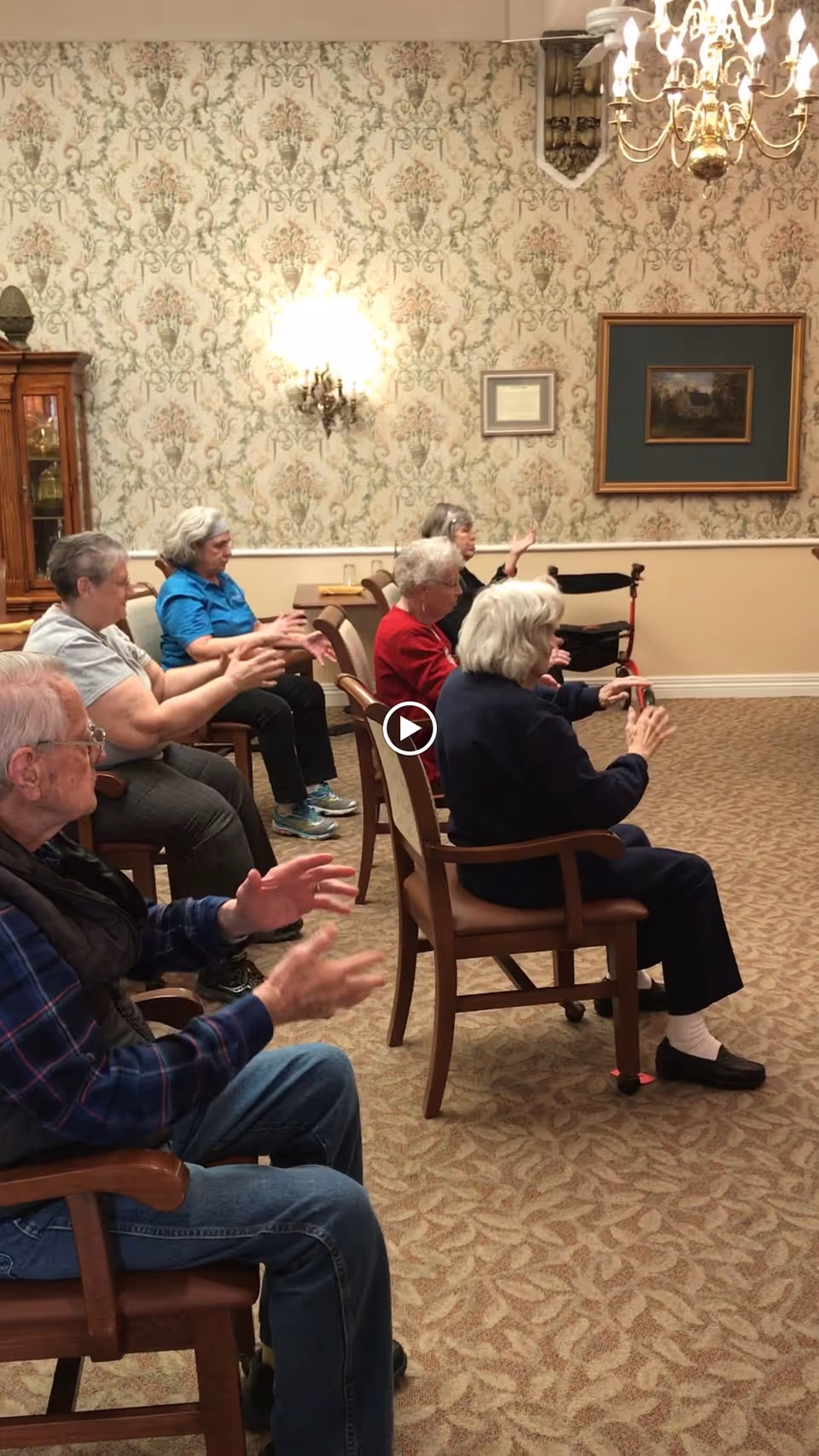 A group of elderly people seated in chairs in a room with floral wallpaper and a chandelier, participating in a seated exercise or activity, with their hands raised and moving.