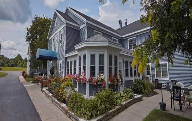 Exterior view of a two-story senior living facility building with blue siding, white trim, and multiple windows. The building features a small garden with colorful flowers and greenery along the sidewalk. There is a blue awning above the entrance and outdoor seating with chairs and tables on the right side. Trees and a partly cloudy sky are visible in the background.