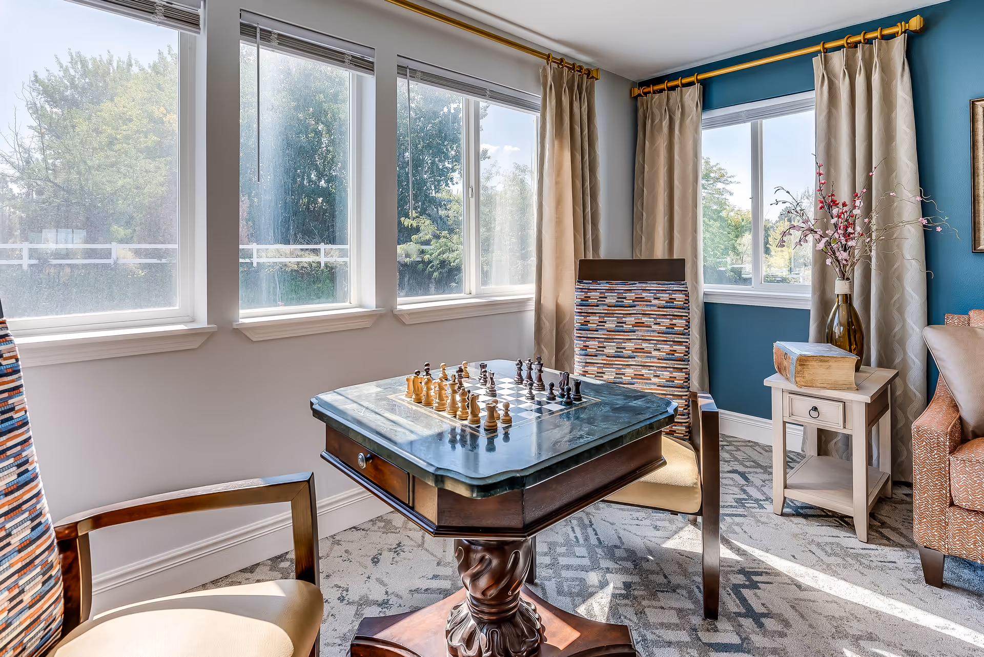 Sunlit sitting area with a chess table and two upholstered chairs beside large windows and a side table with a vase.