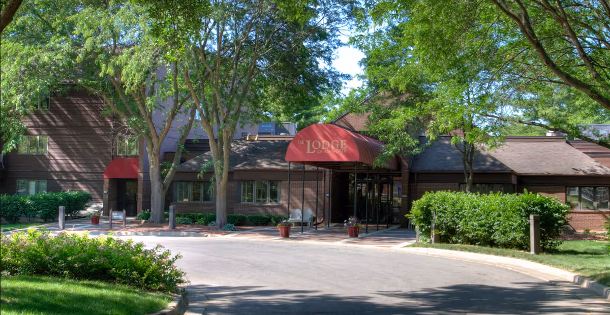 Exterior view of a senior living facility named The Lodge of Ashworth, showing a brick building with a red canopy over the entrance, surrounded by trees and greenery on a sunny day.