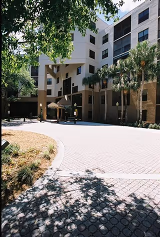 Exterior view of a multi-story senior living facility building with a covered entrance, palm trees, and a paved driveway surrounded by landscaping.