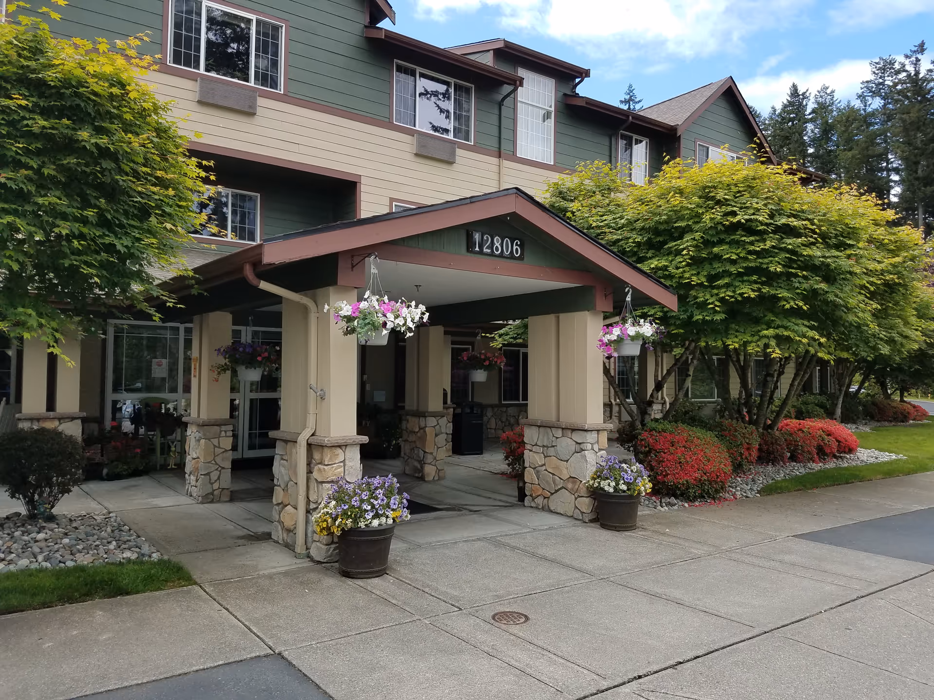Exterior view of a senior living facility entrance with a covered drop-off area supported by stone pillars. The building is green and beige with multiple windows. There are hanging flower pots and flower beds with red and purple flowers, along with well-maintained trees and shrubs around the entrance. The address number 12806 is displayed above the entrance.