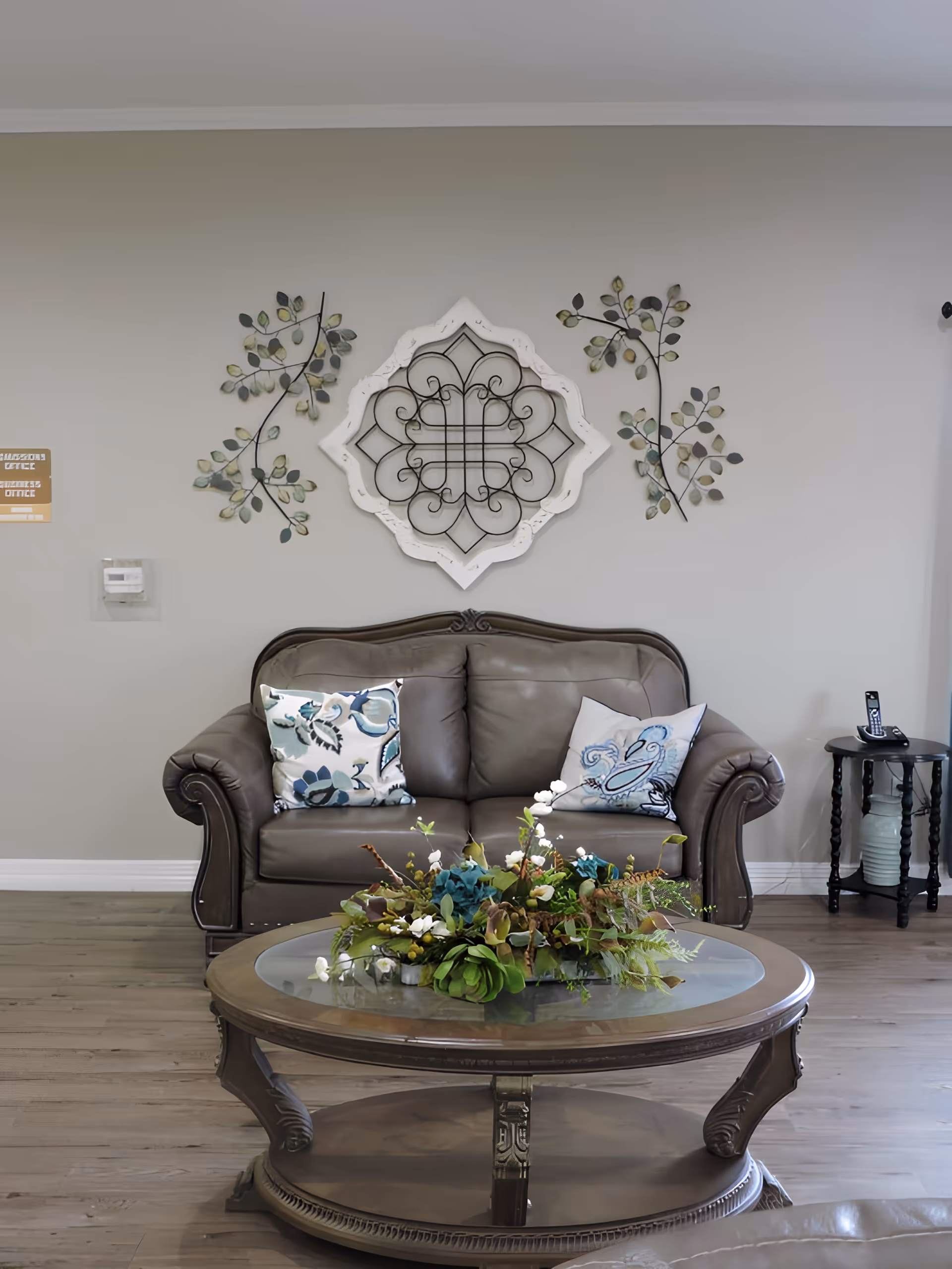 A cozy living room area with a brown leather loveseat adorned with two decorative pillows. In front of the loveseat is a round wooden coffee table with a glass top, featuring a floral arrangement centerpiece. The wall behind the loveseat is decorated with a white ornate frame and two metal leaf wall art pieces. To the right of the loveseat is a small round side table with a cordless phone on it. The room has light-colored wooden flooring and neutral-colored walls.