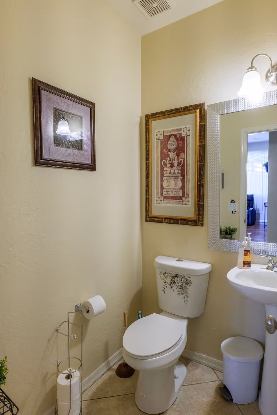 A small bathroom with a white toilet decorated with floral designs, a white pedestal sink with a soap dispenser, a trash can, a toilet paper holder with extra rolls, a plunger, and two framed pictures on the walls. A mirror above the sink reflects part of an adjacent room.