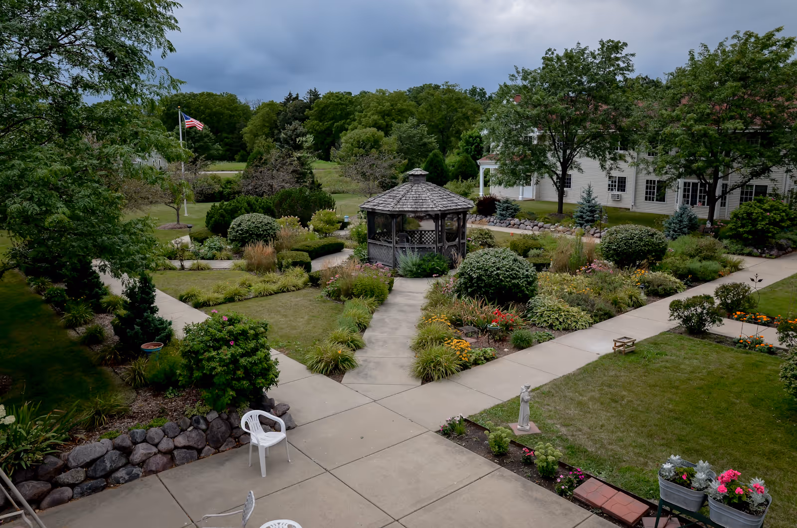 A well-maintained garden area with a central gazebo surrounded by various shrubs, flowers, and trees. Concrete pathways lead to and from the gazebo, and there are white plastic chairs placed along the paths. In the background, there is a two-story building partially visible with windows and trees around it. An American flag is flying on a flagpole to the left side of the garden.