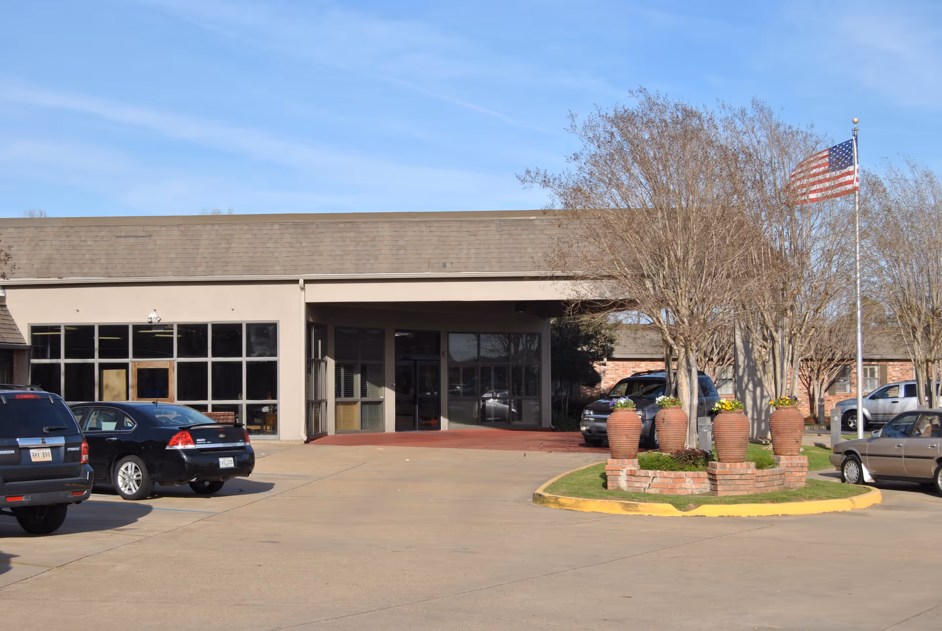 Exterior view of the entrance to Summit Retirement Center with a driveway, parked cars, an American flag on a flagpole, and large decorative planters with flowers and leafless trees.