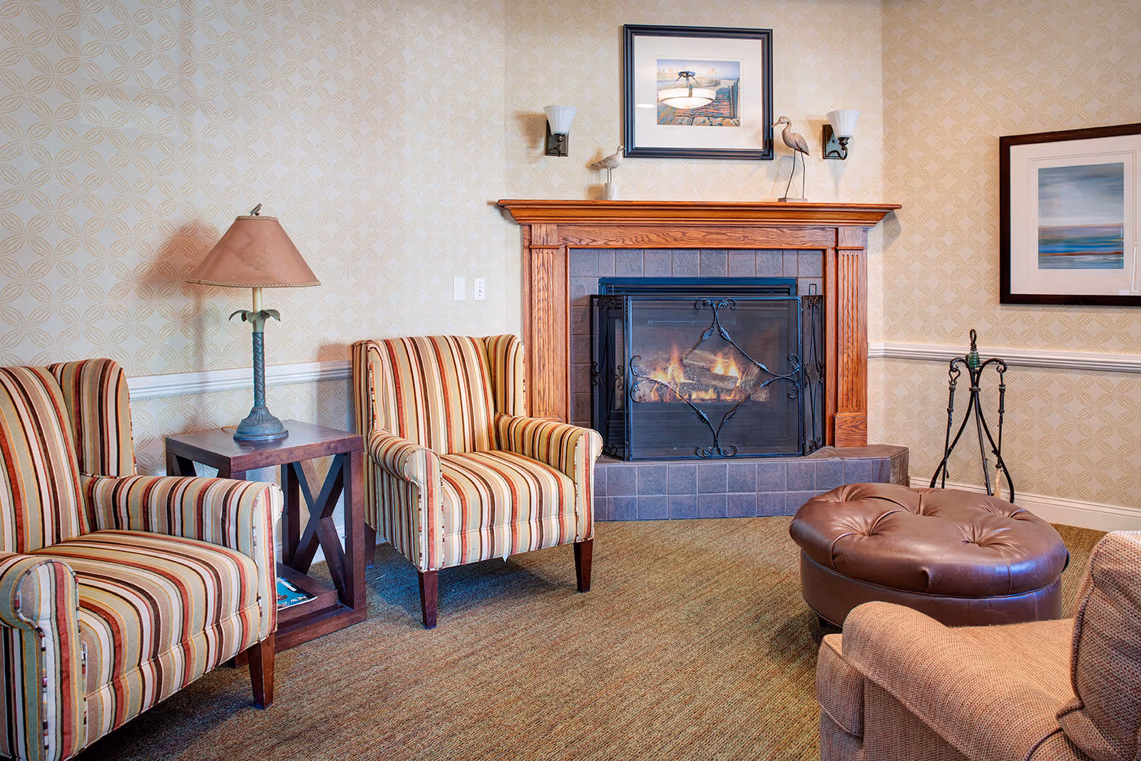 A cozy living room with two striped armchairs, a wooden side table with a lamp, a fireplace with a wooden mantel and a fire burning inside, a round brown leather ottoman, and framed artwork on the walls.