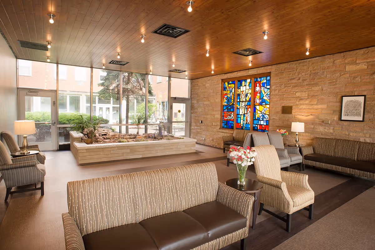 A spacious and well-lit living room area with multiple upholstered chairs and sofas arranged around small tables. The room features a wooden ceiling with recessed lighting, a stone accent wall with a colorful stained glass window, and large floor-to-ceiling windows that provide a view of an outdoor garden area with greenery and a water feature.